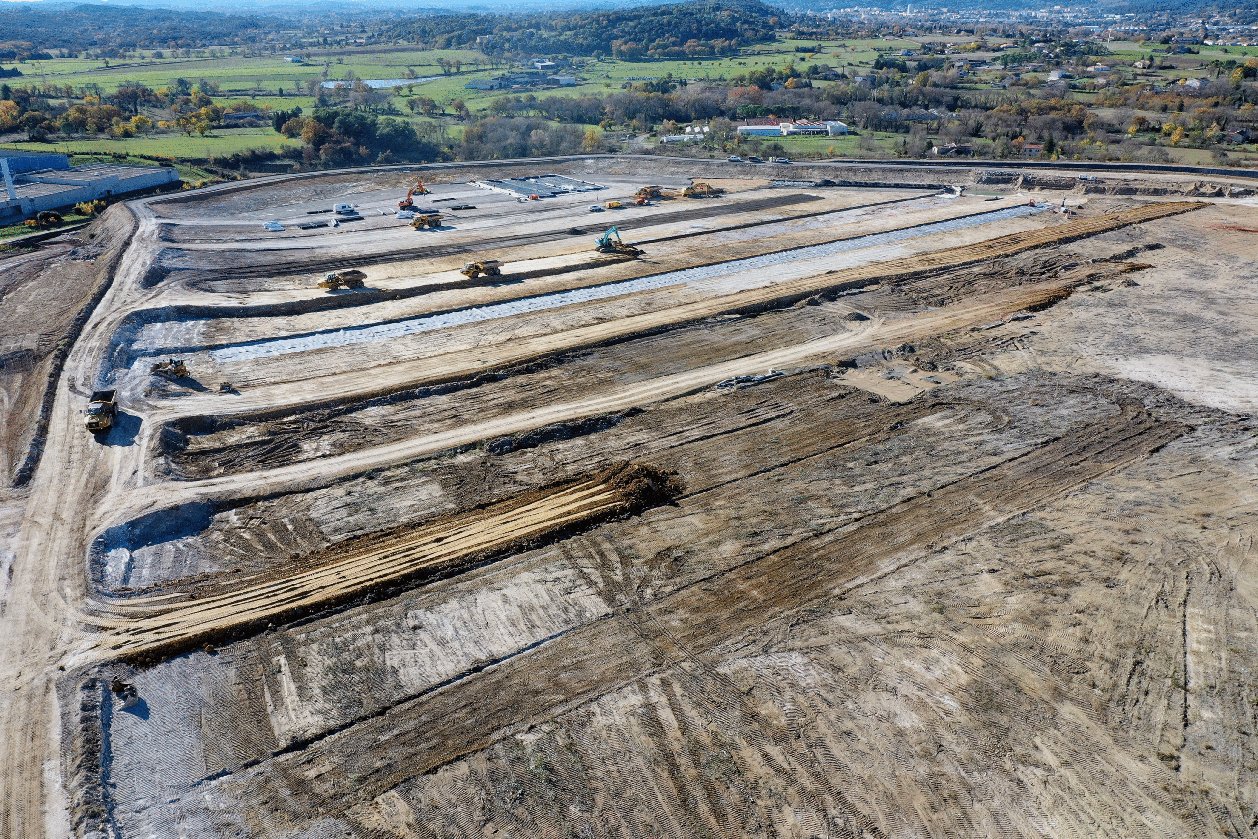 Cette image représente un vaste site industriel en cours de dépollution par Novacis, entreprise spécialisée dans la remédiation industrielle, le suivi environnemental, l'expertise technique, la topographie et la modélisation 3D. La photo prise par drone représente une opération de dépollution de grande envergure, de nombreux véhicules de chantier qui excavent et transportent la terre polluée