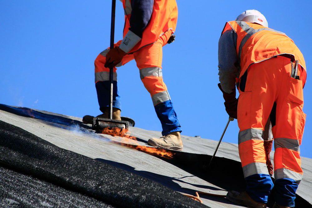 Deux ouvriers en tenue de sécurité posent une membrane étanche sur portion de chantier inclinée, utilisant une technique de soudure à chaud